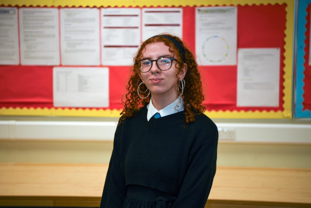A person with curly red hair, glasses, and hoop earrings stands in a classroom in front of a bulletin board displaying various papers and charts. They wear a dark sweater over a collared shirt and look toward the camera.
