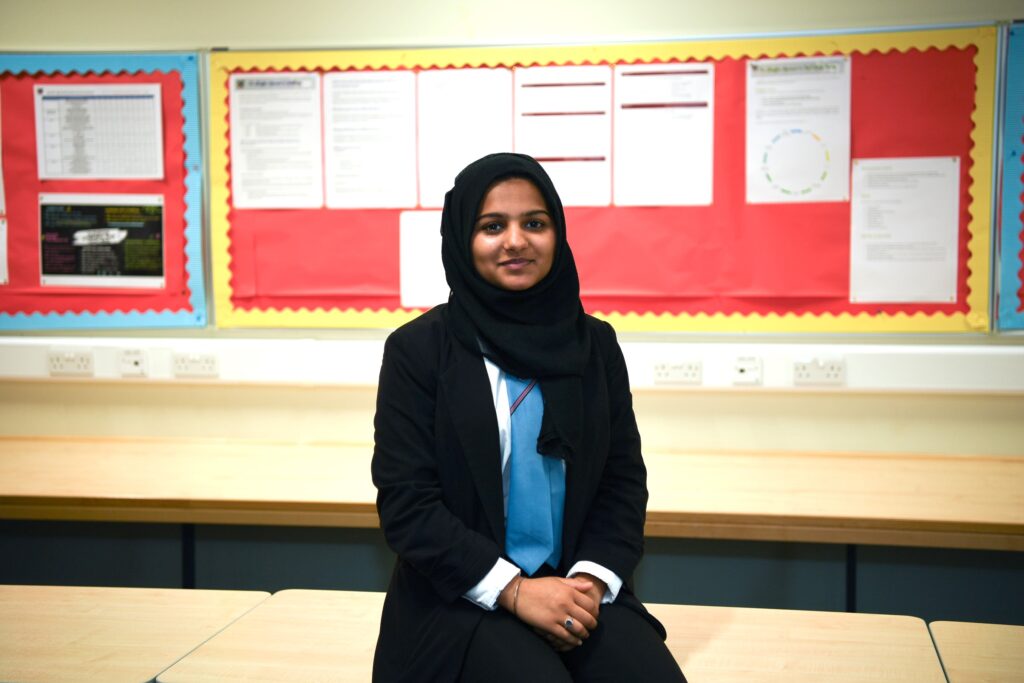 A woman wearing a black hijab and suit sits on a desk in a classroom, smiling slightly. Behind her are red bulletin boards with various papers and notices pinned up.
