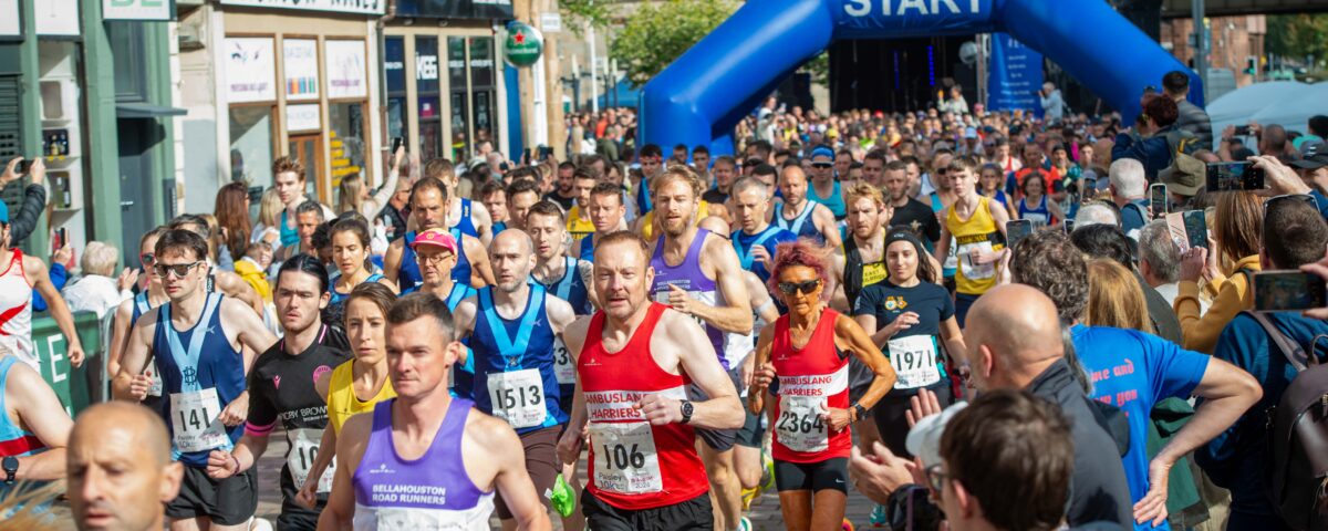 A large group of runners in colorful athletic gear start a race on a city street, passing under a blue inflatable arch labeled "START," with spectators lining the sides and buildings in the background.