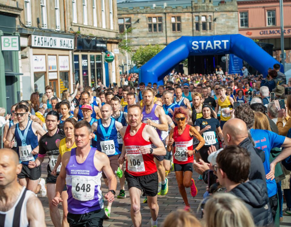 A large group of runners in colorful athletic gear start a race on a city street, passing under a blue inflatable arch labeled "START," with spectators lining the sides and buildings in the background.