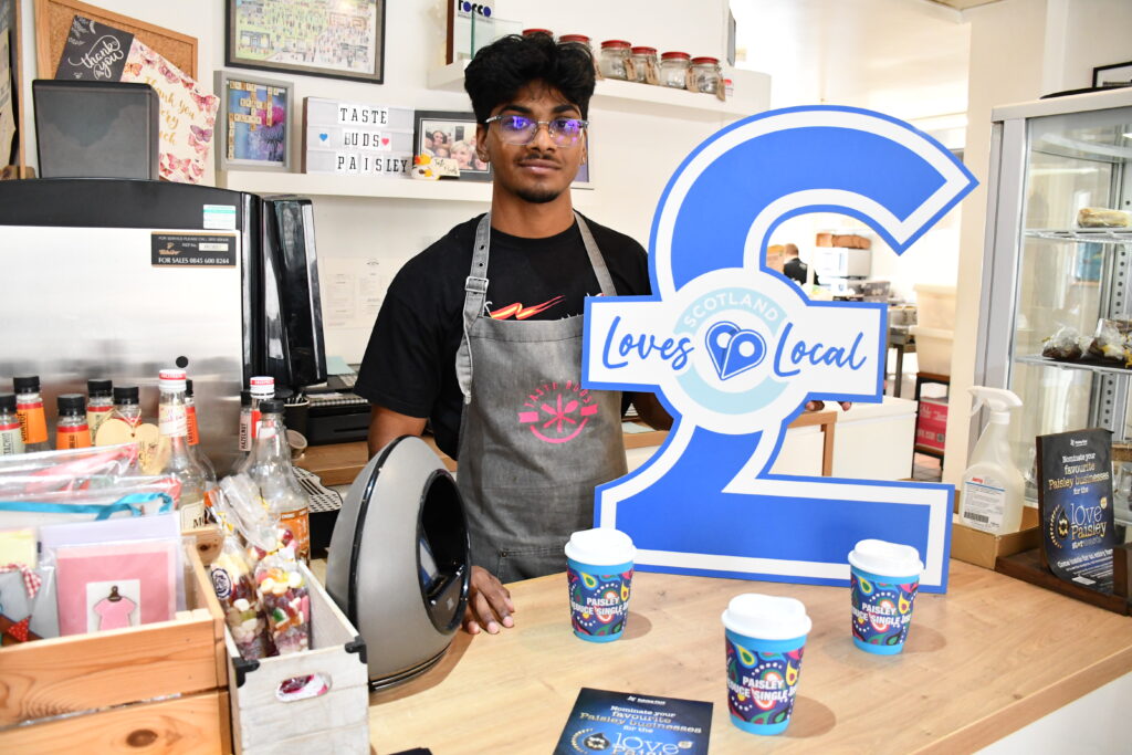 A young man stands behind a café counter holding a large blue "Scotland Loves Local" pound sign, surrounded by snacks, colorful cups, and promotional materials in a bright shop.