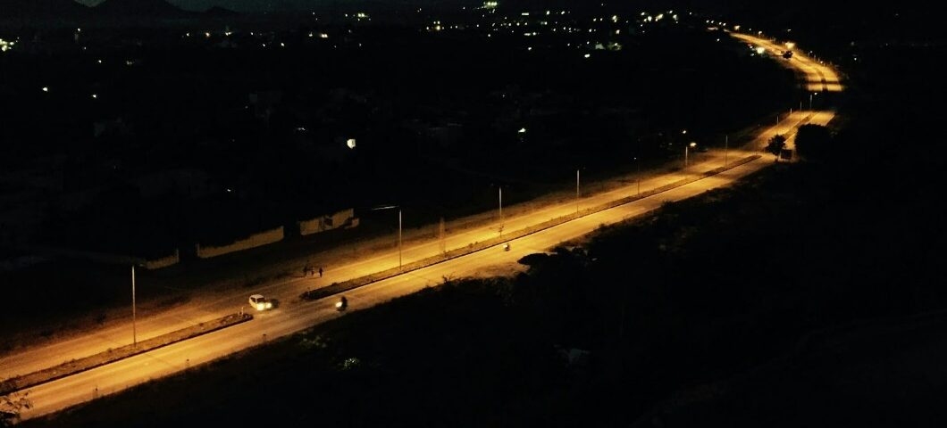 Aerial view of a road at night, illuminated by streetlights. The road curves through a dark landscape with scattered lights in the distance and silhouettes of hills against the evening sky.