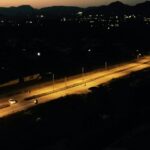 Aerial view of a road at night, illuminated by streetlights. The road curves through a dark landscape with scattered lights in the distance and silhouettes of hills against the evening sky.