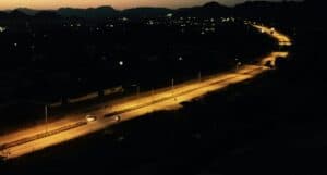 Aerial view of a road at night, illuminated by streetlights. The road curves through a dark landscape with scattered lights in the distance and silhouettes of hills against the evening sky.