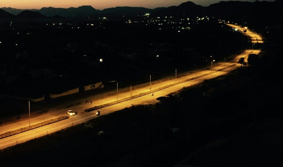 Aerial view of a road at night, illuminated by streetlights. The road curves through a dark landscape with scattered lights in the distance and silhouettes of hills against the evening sky.