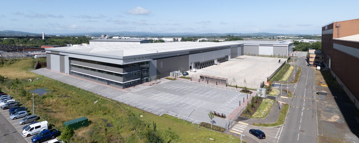 Aerial view of a large, modern warehouse with a wide parking lot, surrounded by grass and a road with parked cars under a clear, blue sky.