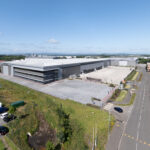 Aerial view of a large, modern warehouse with a wide parking lot, surrounded by grass and a road with parked cars under a clear, blue sky.