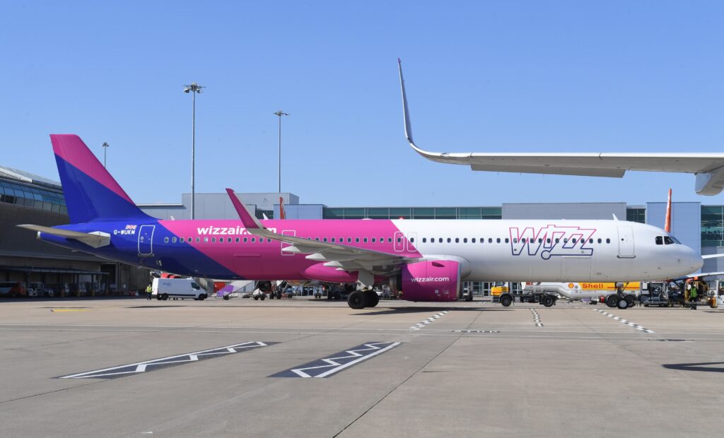 A Wizz Air airplane with a pink and purple livery is parked at an airport gate, connected to a jet bridge, with airport buildings and equipment in the background under a clear blue sky.