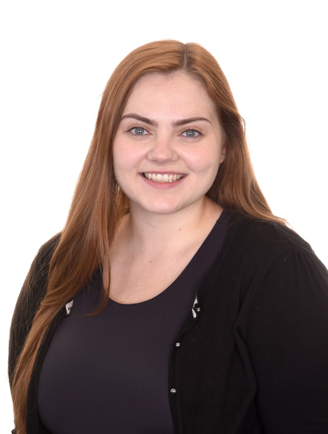 A young woman with long auburn hair and light skin smiles at the camera. She is wearing a dark top and a black cardigan against a plain white background.