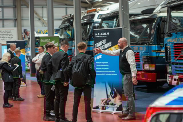 A group of students listens to a man giving a presentation beside several parked trucks indoors, with informational banners displayed behind them.