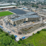 Aerial view of a large construction site featuring a partially built modern building surrounded by dirt roads, machinery, green fields, and nearby industrial and residential areas.