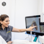 A dentist in a gray uniform smiles and points to a digital 3D dental scan displayed on a computer screen in a bright, modern clinic.
