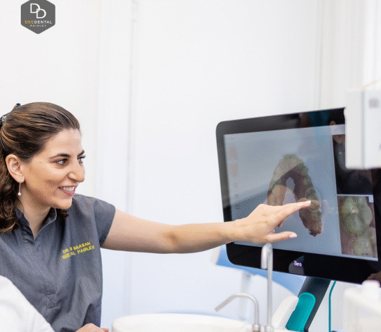 A dentist in a gray uniform smiles and points to a digital 3D dental scan displayed on a computer screen in a bright, modern clinic.