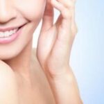Close-up of a smiling woman with clear skin, showing her lower face, shoulder, and hand near her face, against a soft blue background.