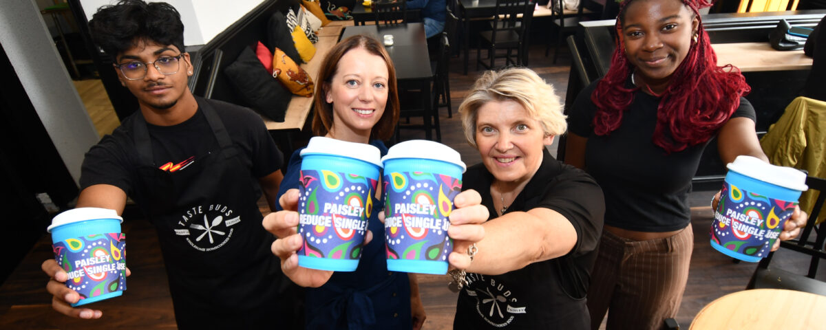 Four people stand in a café, smiling and holding up colorful takeaway coffee cups labeled "Paisley People’s Shuffle," with customers sitting in the background.