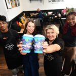 Four people stand in a café, smiling and holding up colorful takeaway coffee cups labeled "Paisley People’s Shuffle," with customers sitting in the background.