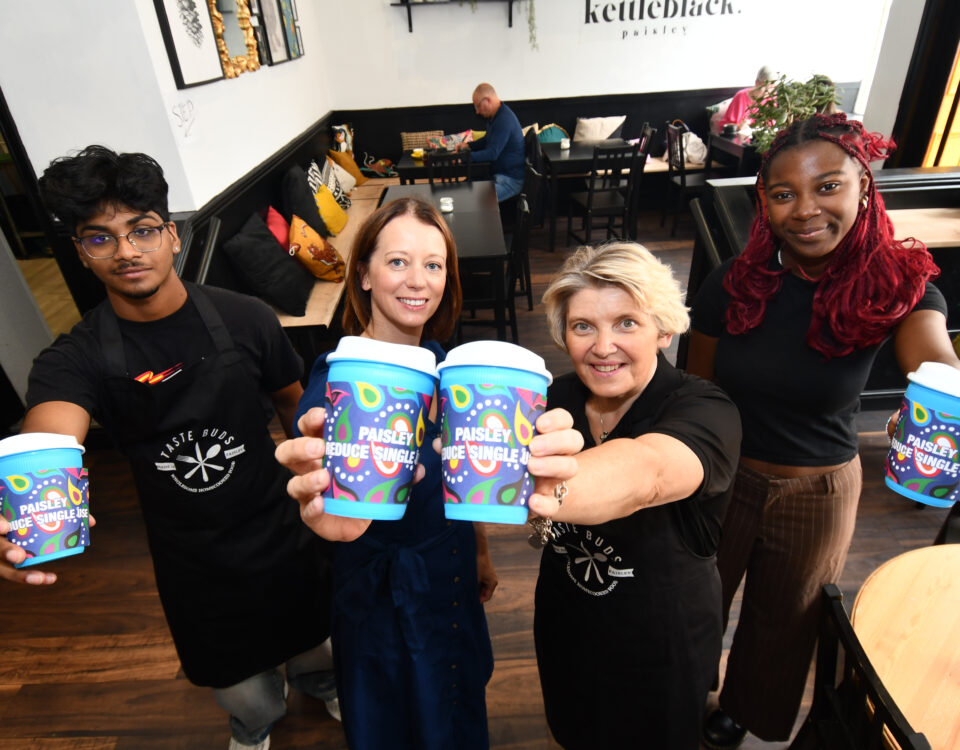 Four people stand in a café, smiling and holding up colorful takeaway coffee cups labeled "Paisley People’s Shuffle," with customers sitting in the background.