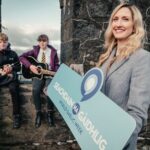 A smiling woman in a gray blazer holds a "Seachdain na Gàidhlig—World Gaelic Week" sign, while two young men sit on a stone wall behind her, one playing a guitar and the other with a mandolin.