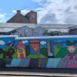 A colorful mural on a long wall features playful drawings of children, animals, houses, nature, and the word "WILLYBUGS" in bold letters, with industrial buildings and a partly cloudy sky in the background.