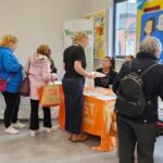 People interact at information tables inside a community center. Some are standing, talking, or receiving pamphlets, while others sit behind tables with flyers and displays. A snack table is visible to the left.