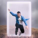 A man in a blue shirt and black pants appears to leap energetically through a glowing, rectangular portal, with cloudy skies and grassy terrain in the background.