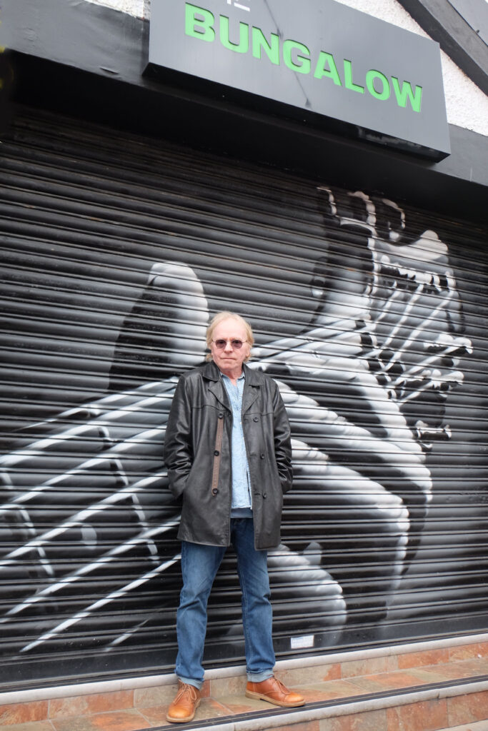 A man in sunglasses and a black leather jacket stands in front of a shutter with a large black-and-white mural of a hand playing a guitar. The sign above him reads "BUNGALOW" in green letters.