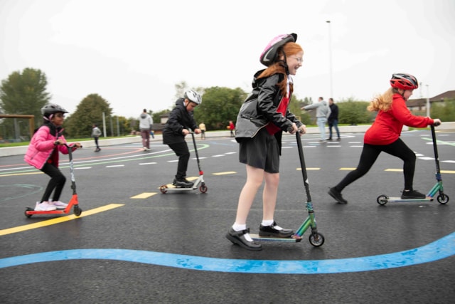 Four children wearing helmets ride scooters on a playground with colorful markings. The kids appear to be having fun, and the weather looks overcast. Trees and playground equipment are visible in the background.