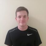 A young man with short brown hair wearing a black Nike T-shirt stands in front of a plain light-colored wall, looking at the camera and smiling slightly.