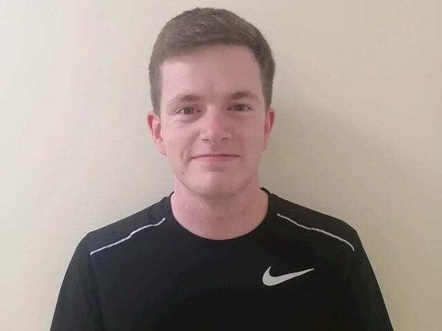 A young man with short brown hair wearing a black Nike T-shirt stands in front of a plain light-colored wall, looking at the camera and smiling slightly.