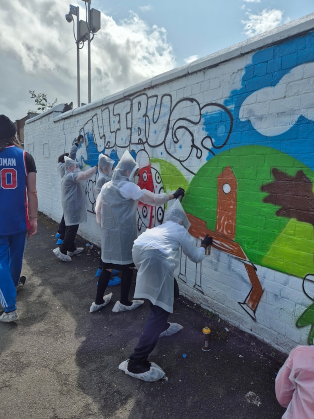 Several people wearing plastic ponchos and shoe covers are spray painting a colorful mural with a horse and blue sky on a white brick wall outdoors. A person in a blue jersey stands nearby.