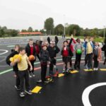 A group of children stands and plays together on an outdoor playground, some holding balls and one with a scooter, while a few jump in the air. The playground has various colorful lines and markings.
