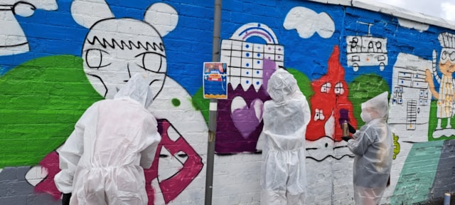 Three people in white coveralls paint a colorful mural featuring cartoon-like characters and buildings on a brick wall. One person holds a phone, likely taking a photo, while the others paint the mural.
