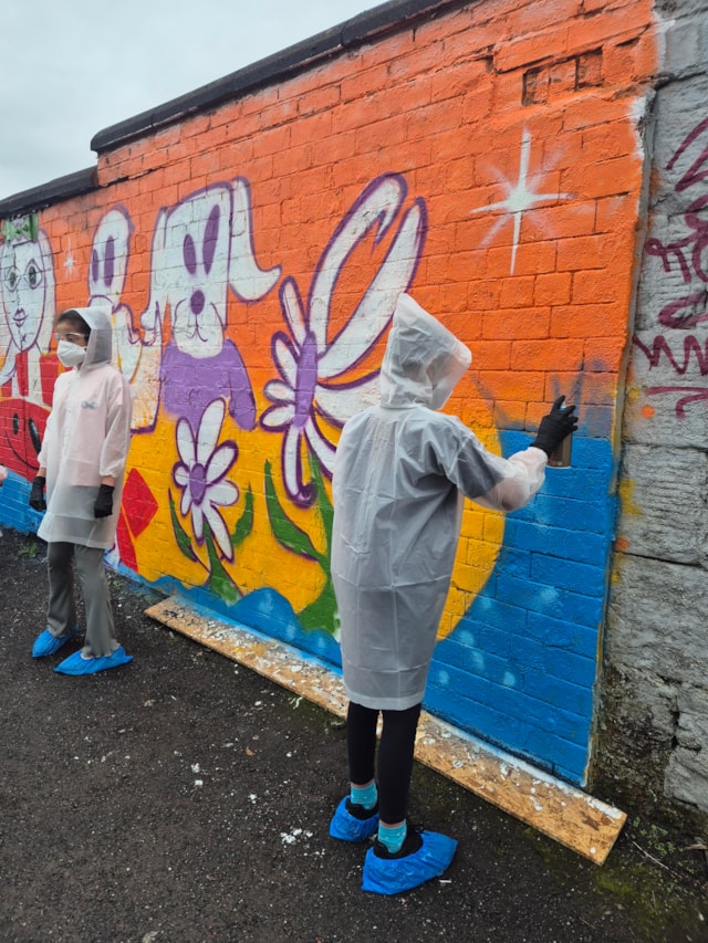 Two people wearing rain ponchos, masks, gloves, and blue shoe covers paint a colorful mural of animals, flowers, and stars on a brick wall outdoors under a cloudy sky.