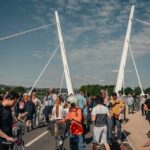 A crowd of people, some with bicycles, gather on a modern white suspension bridge under a partly cloudy sky during the day. Trees and buildings are visible in the background.