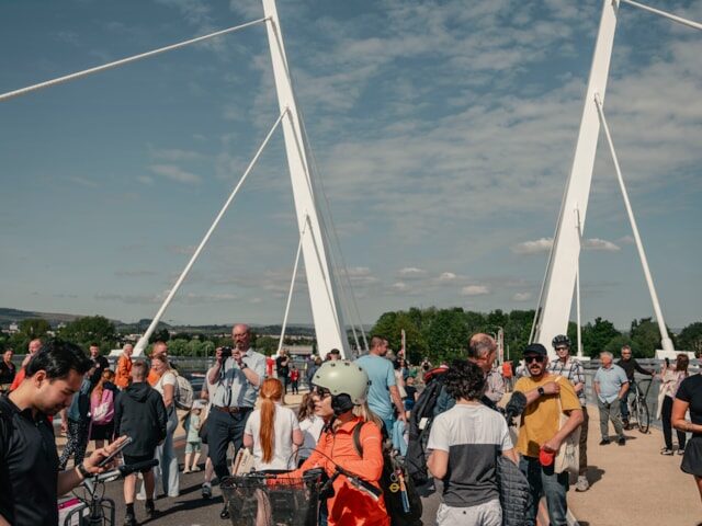 A crowd of people, some with bicycles, gather on a modern white suspension bridge under a partly cloudy sky during the day. Trees and buildings are visible in the background.