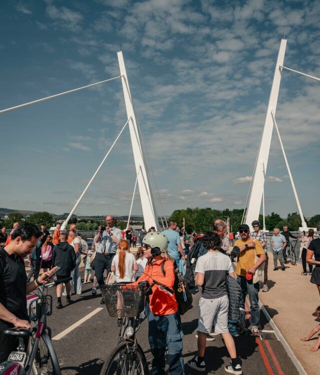 A crowd of people, some with bicycles, gather on a modern white suspension bridge under a partly cloudy sky during the day. Trees and buildings are visible in the background.