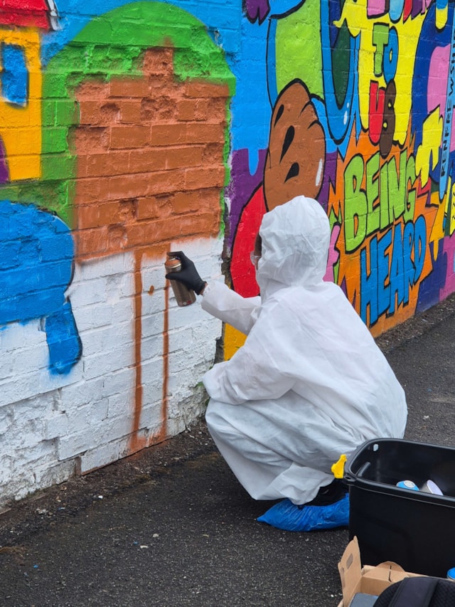 A person in a white protective suit and blue shoe covers spray paints a brick design onto a colorful mural on a brick wall, with paint supplies in a black bin nearby.