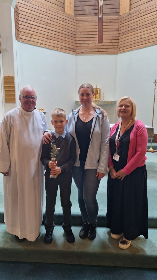 Four people stand together in a church: a man in white robes, a boy in a suit holding a trophy, and two women in casual clothing, all smiling. A cross and altar are visible in the background.