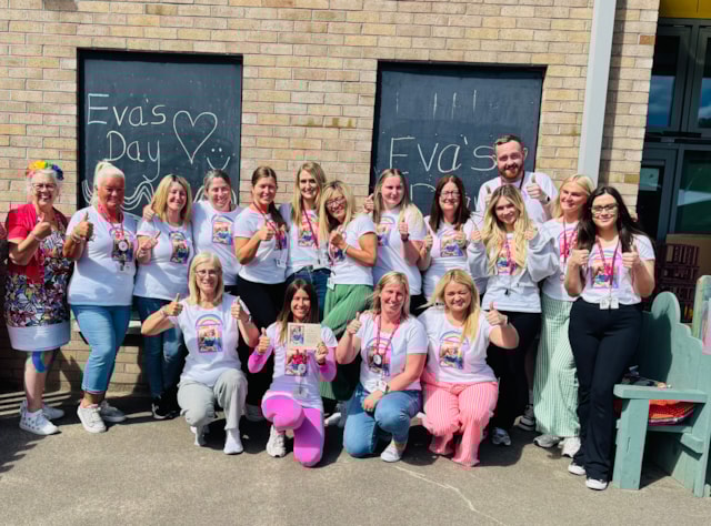 A group of people pose and smile outside in front of two chalkboards reading “Eva’s Day” with hearts. Most wear matching T-shirts with a rainbow design, and some hold up printed sheets featuring a young girl's photo.