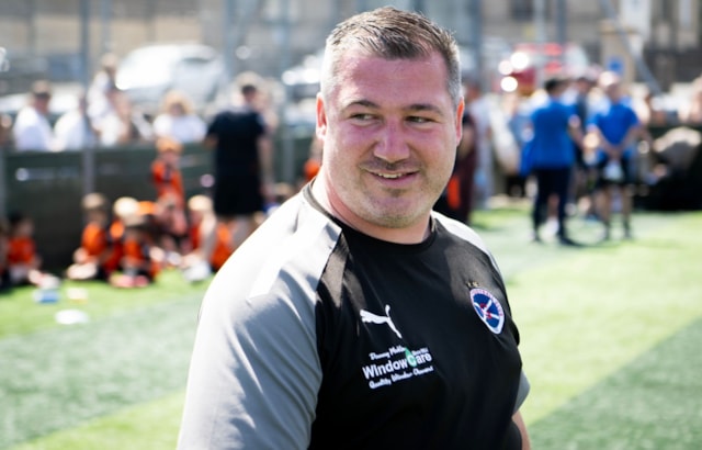 A smiling man in a sports shirt stands on a grassy field with blurred people, some wearing orange, in the background. The scene appears to be at an outdoor sports event on a sunny day.
