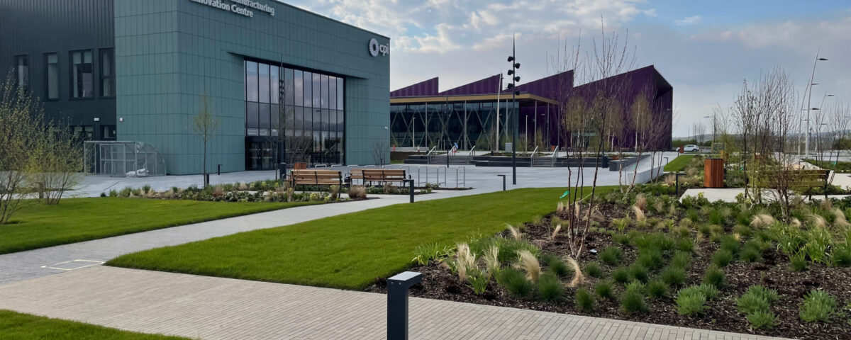 Modern building labeled "Medicines Manufacturing Innovation Centre" with large windows and a landscaped garden featuring green lawns, walkways, benches, small trees, and ornamental grasses under a partly cloudy sky.