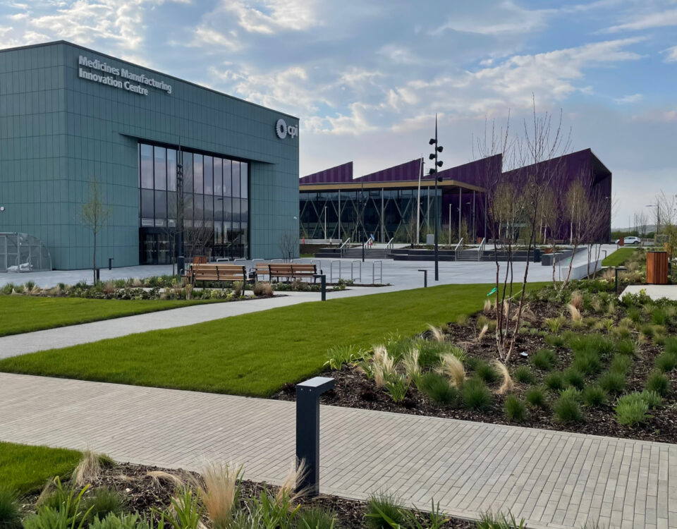 Modern building labeled "Medicines Manufacturing Innovation Centre" with large windows and a landscaped garden featuring green lawns, walkways, benches, small trees, and ornamental grasses under a partly cloudy sky.