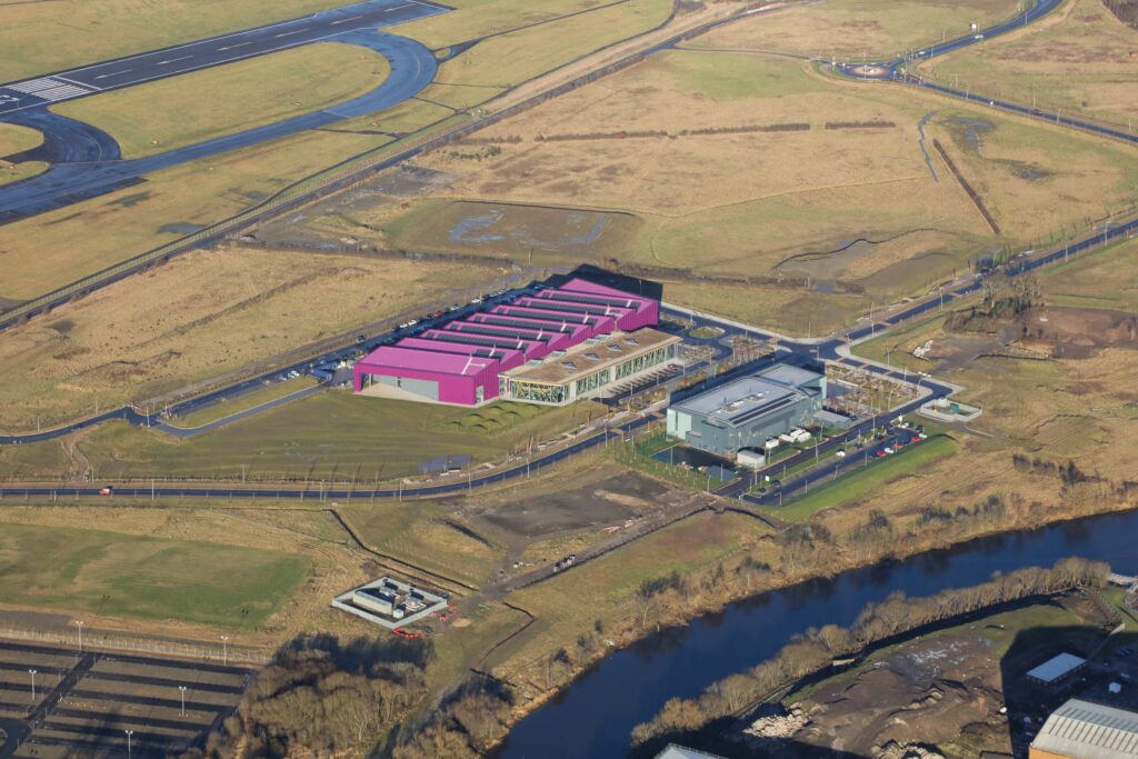Aerial view of a large industrial facility with pink roofs, surrounded by fields, a river, and a nearby road. The site includes multiple buildings and parking areas, alongside a fenced perimeter.