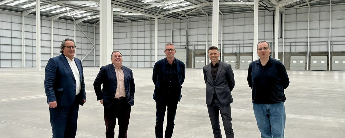 Five men in business and casual attire stand spaced apart in a large, empty warehouse with high ceilings and skylights. The space is open, with metal beams and closed loading doors visible in the background.