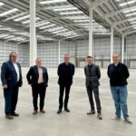 Five men in business and casual attire stand spaced apart in a large, empty warehouse with high ceilings and skylights. The space is open, with metal beams and closed loading doors visible in the background.
