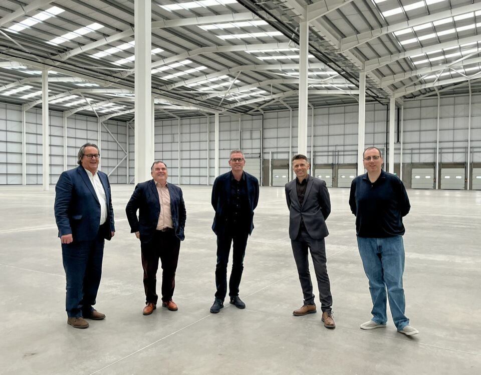 Five men in business and casual attire stand spaced apart in a large, empty warehouse with high ceilings and skylights. The space is open, with metal beams and closed loading doors visible in the background.