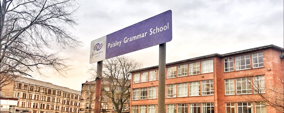 A purple sign reading "Paisley Grammar School" stands in front of a red brick school building and a parking lot filled with cars under a cloudy sky.