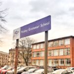 A purple sign reading "Paisley Grammar School" stands in front of a red brick school building and a parking lot filled with cars under a cloudy sky.