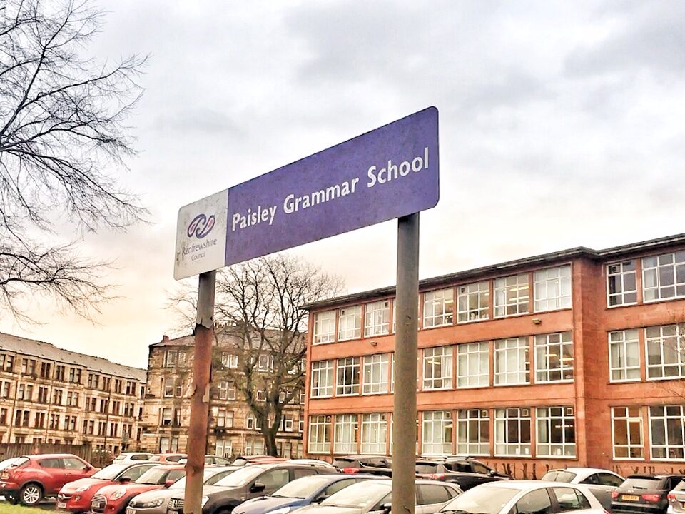 A purple sign reading "Paisley Grammar School" stands in front of a red brick school building and a parking lot filled with cars under a cloudy sky.