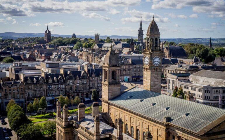 A cityscape with historic stone buildings, a prominent clock tower in the foreground, and lush green hills in the background under a partly cloudy sky.